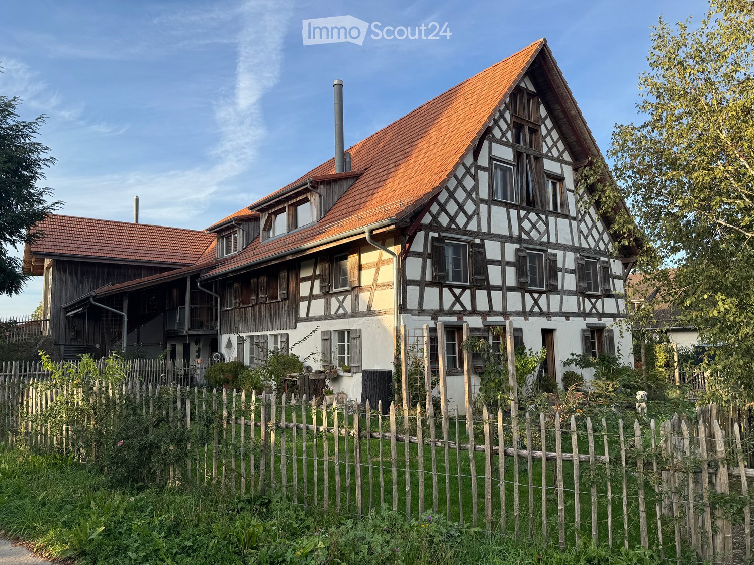 Traditional half-timbered house with red-tiled roof, white walls, multiple windows, and a wooden fence surrounding the garden.