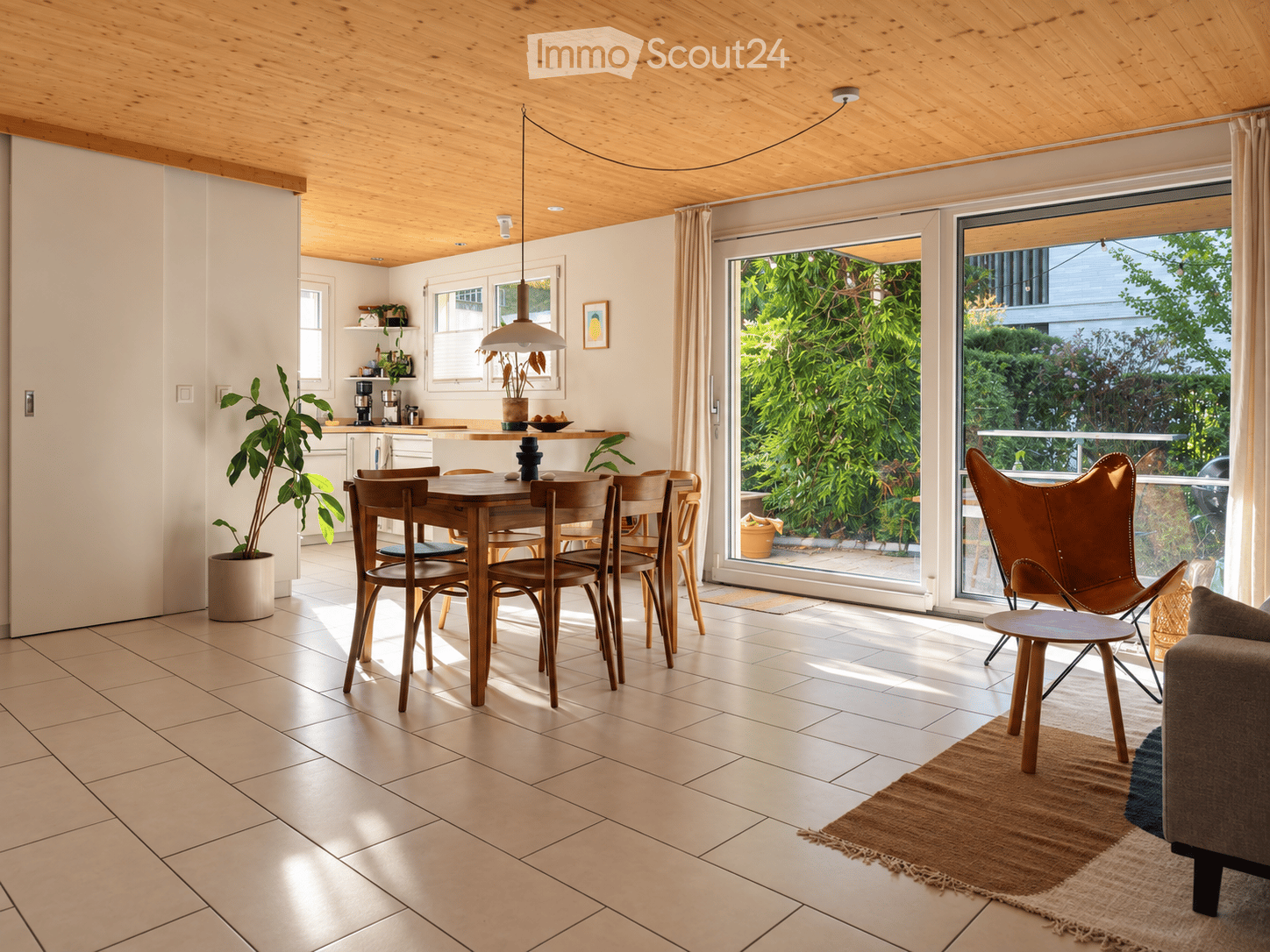 Bright, open-plan living and dining area with wooden dining table, chairs, sofa, pendant light, and large windows overlooking a garden.