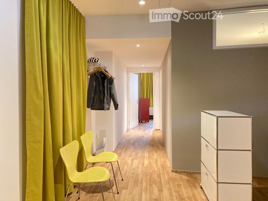 A narrow hallway with wooden flooring, two bright yellow chairs, a white cabinet, and a coat rack with jackets hanging on it. A doorway leads to another room with a red wardrobe and yellow curtains.