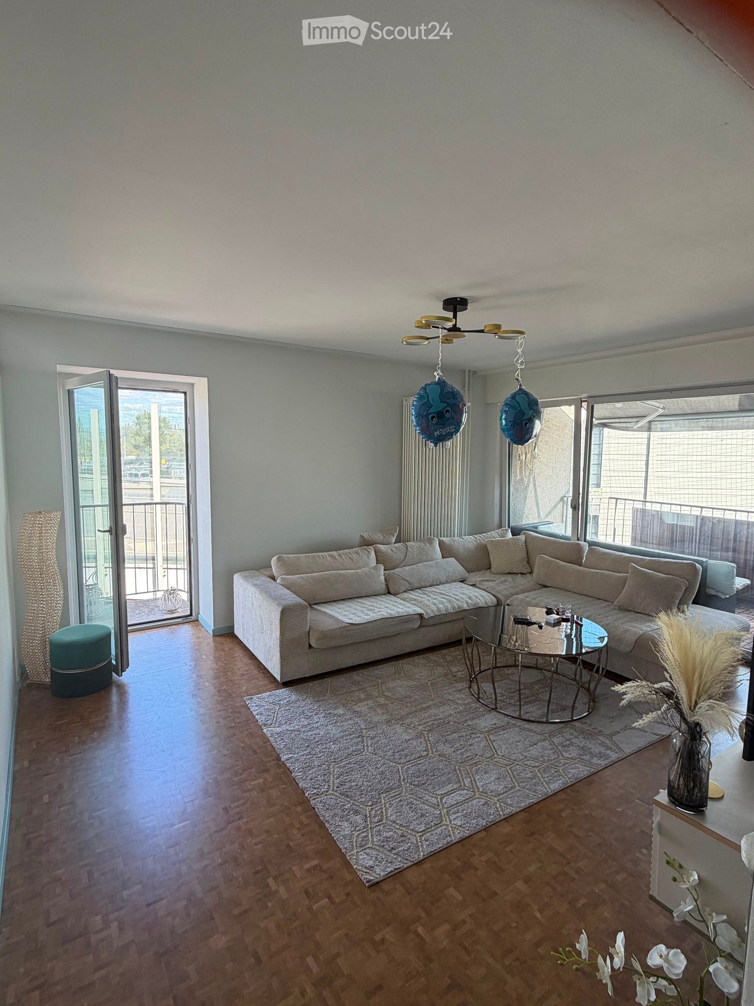 Modern living room with a large sectional sofa, coffee table, round glass table, and decorative vases. Features a balcony with a view and a hanging light fixture.