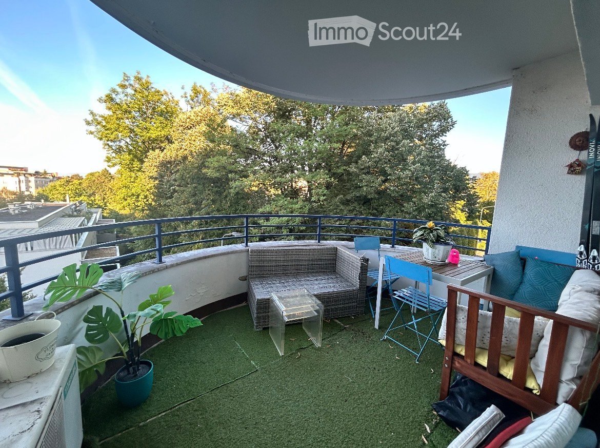 Balcony with wicker sofa, small table, chairs, potted plants, and a view of trees and buildings.