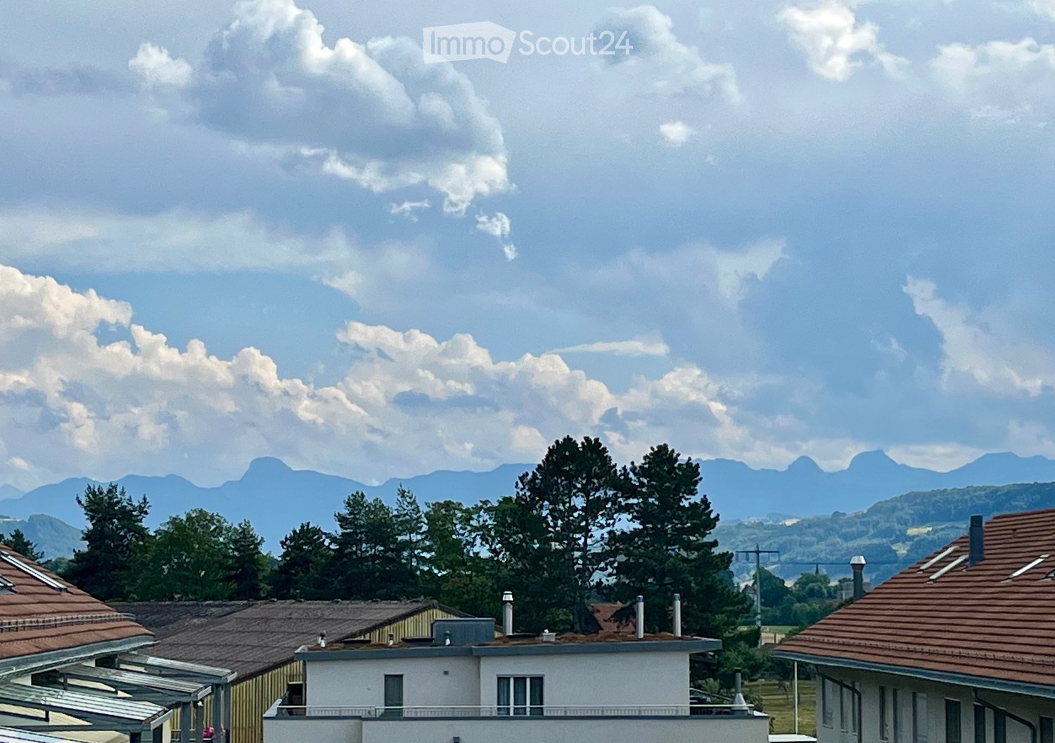 Residential buildings with tiled roofs, surrounded by trees and distant mountains under a partly cloudy sky.