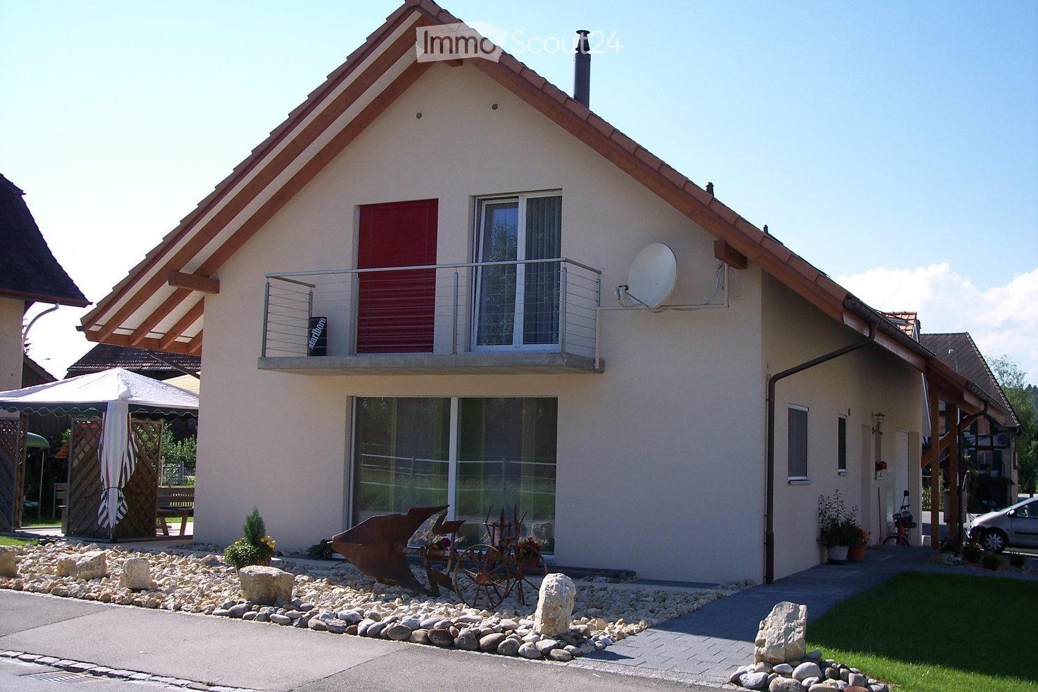 Two-story house with a beige exterior, red shutters, and a balcony on the upper level. Features include a satellite dish, a small garden area with decorative rocks and plants, and a covered patio with outdoor furniture.