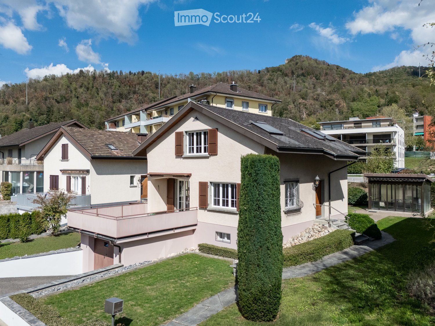 Two-story house with beige walls, brown shutters, and a tiled roof, situated in a green, hilly area with neighboring houses and a scenic backdrop.