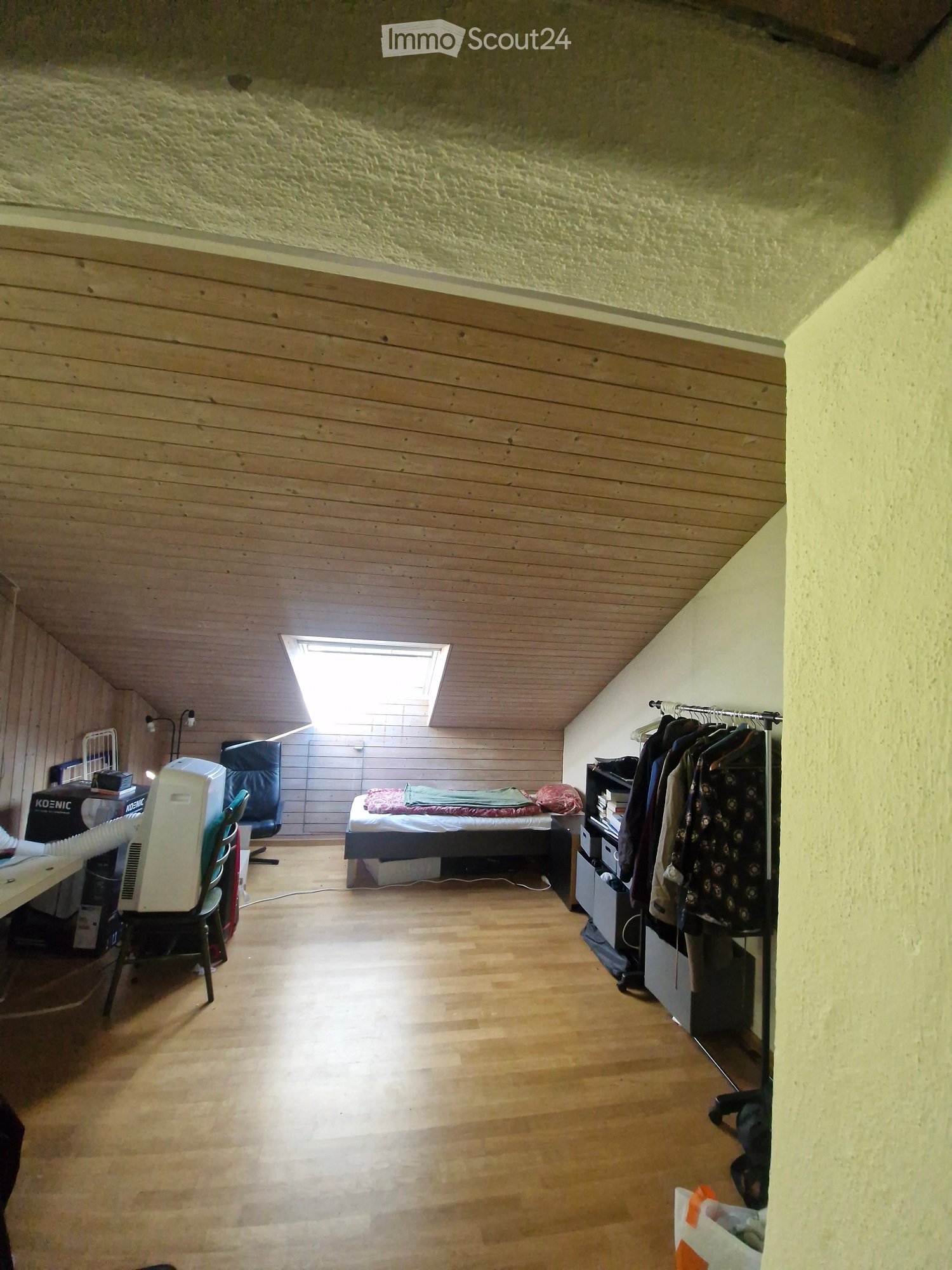 A furnished bedroom with wooden flooring, a bed, a desk, a chair, a wardrobe, and an air conditioner. The room has a slanted wooden ceiling and a skylight window.