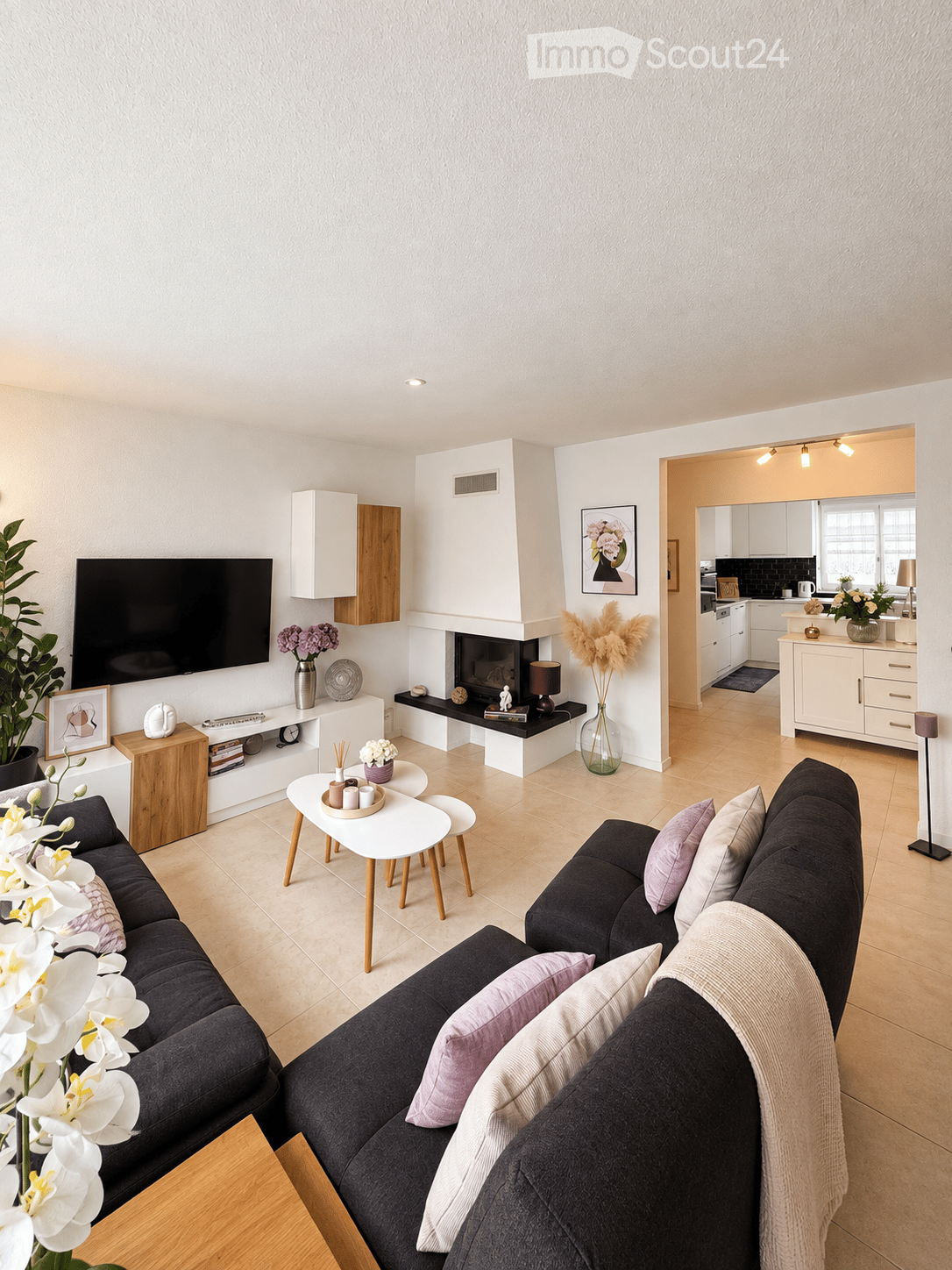 Modern living room with a dark grey sectional sofa, white coffee table, fireplace, flat-screen TV, and decorative plants.
