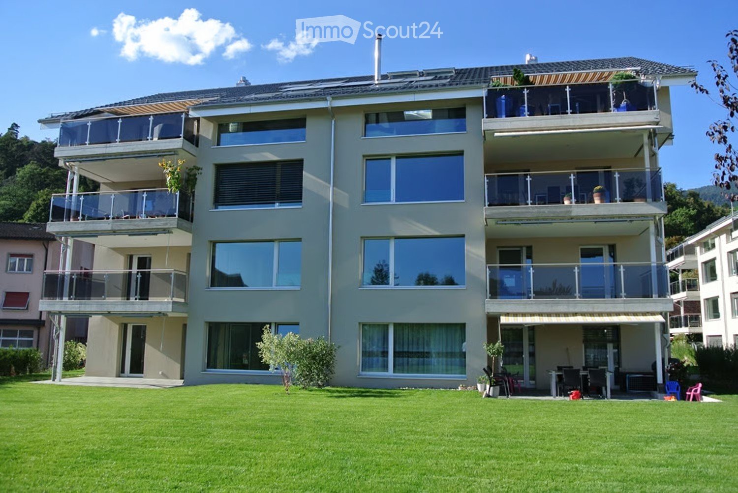 Modern three-story house with large windows and multiple balconies, surrounded by a well-maintained lawn and greenery.