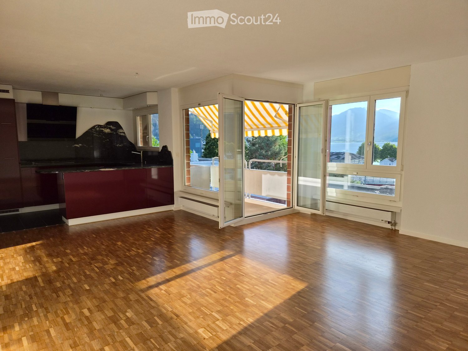 Bright, unfurnished room with wooden flooring, large windows, and a view of mountains and a lake. The room features a modern kitchen area with dark cabinets and a countertop, and large glass doors open to a balcony with striped awnings.