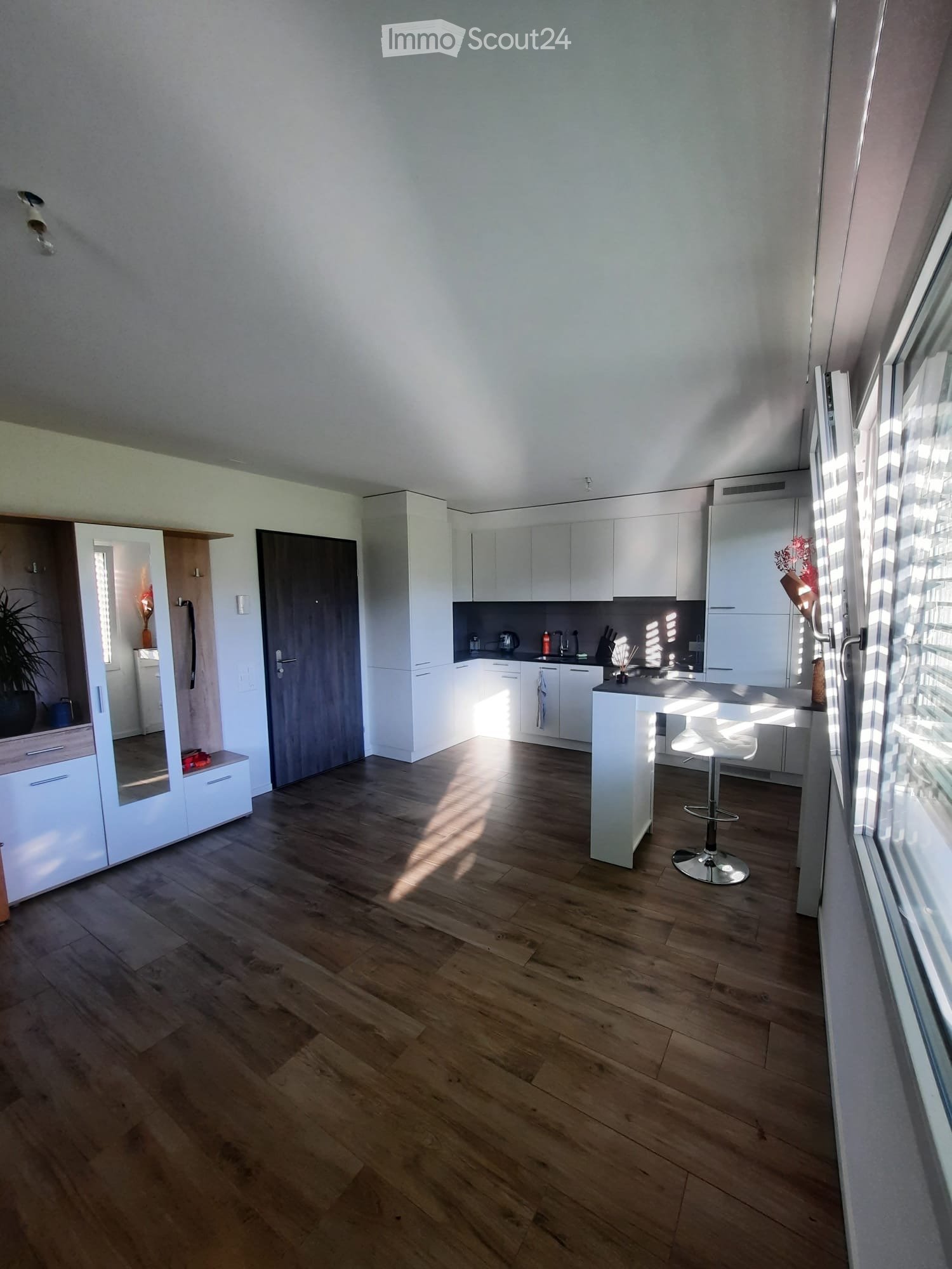 Modern kitchen with white cabinets, wooden floor, island with stool, and large windows letting in natural light.