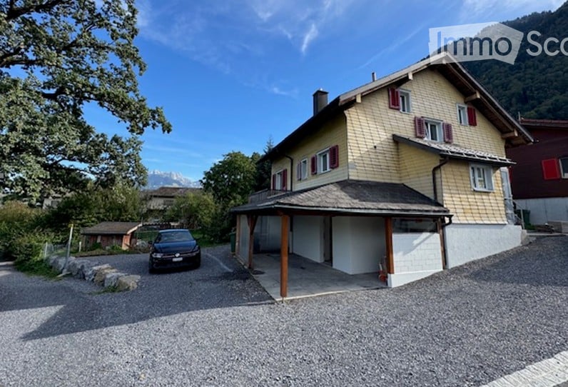 Two-story house with yellow siding, red shutters, and a covered garage. A car is parked in the driveway, and the house is situated in a scenic area with mountains in the background.