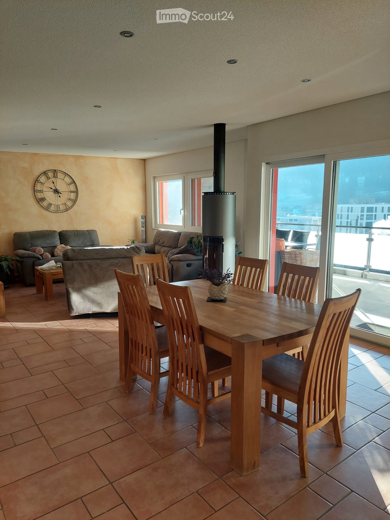 Spacious living and dining area with tiled flooring, large windows, and a balcony. Features a sofa, dining table with chairs, a clock, and a wood-burning stove.