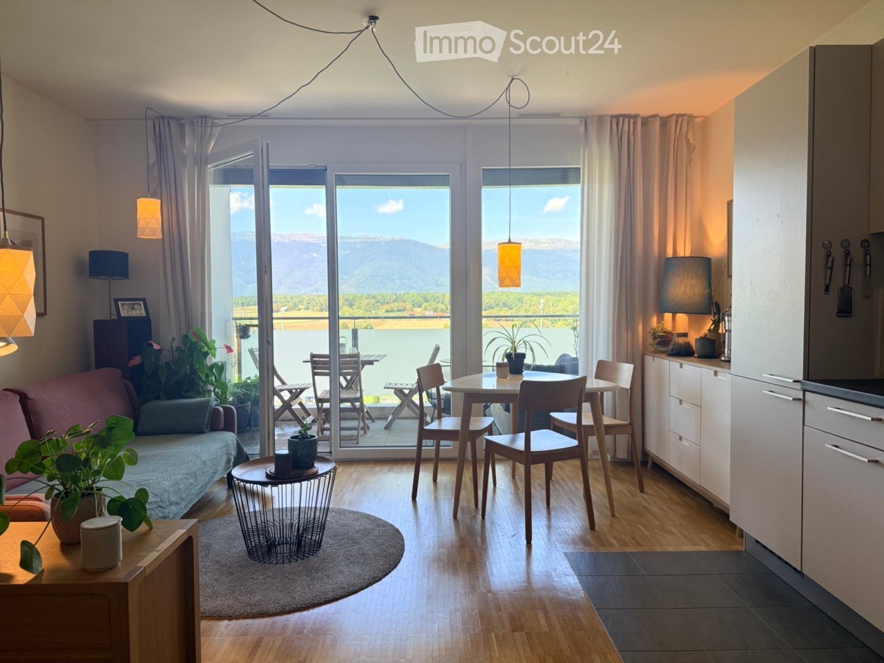 Bright, open-plan living and dining area with large windows overlooking a scenic lake and mountains. Features a sofa, coffee table, dining table with chairs, kitchen area with cabinets and appliances, pendant lights, and decorative plants.