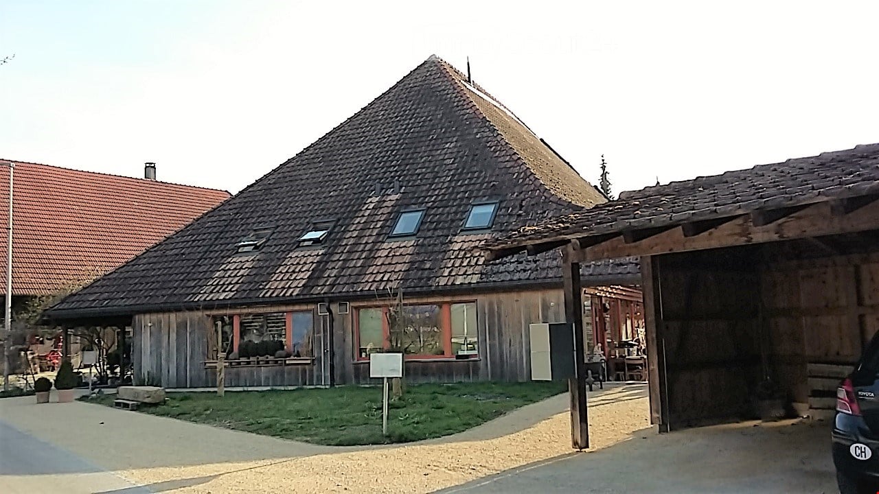 Traditional wooden building with a steep, tiled roof and a covered garage area. The structure features large windows and a small grassy area in front.