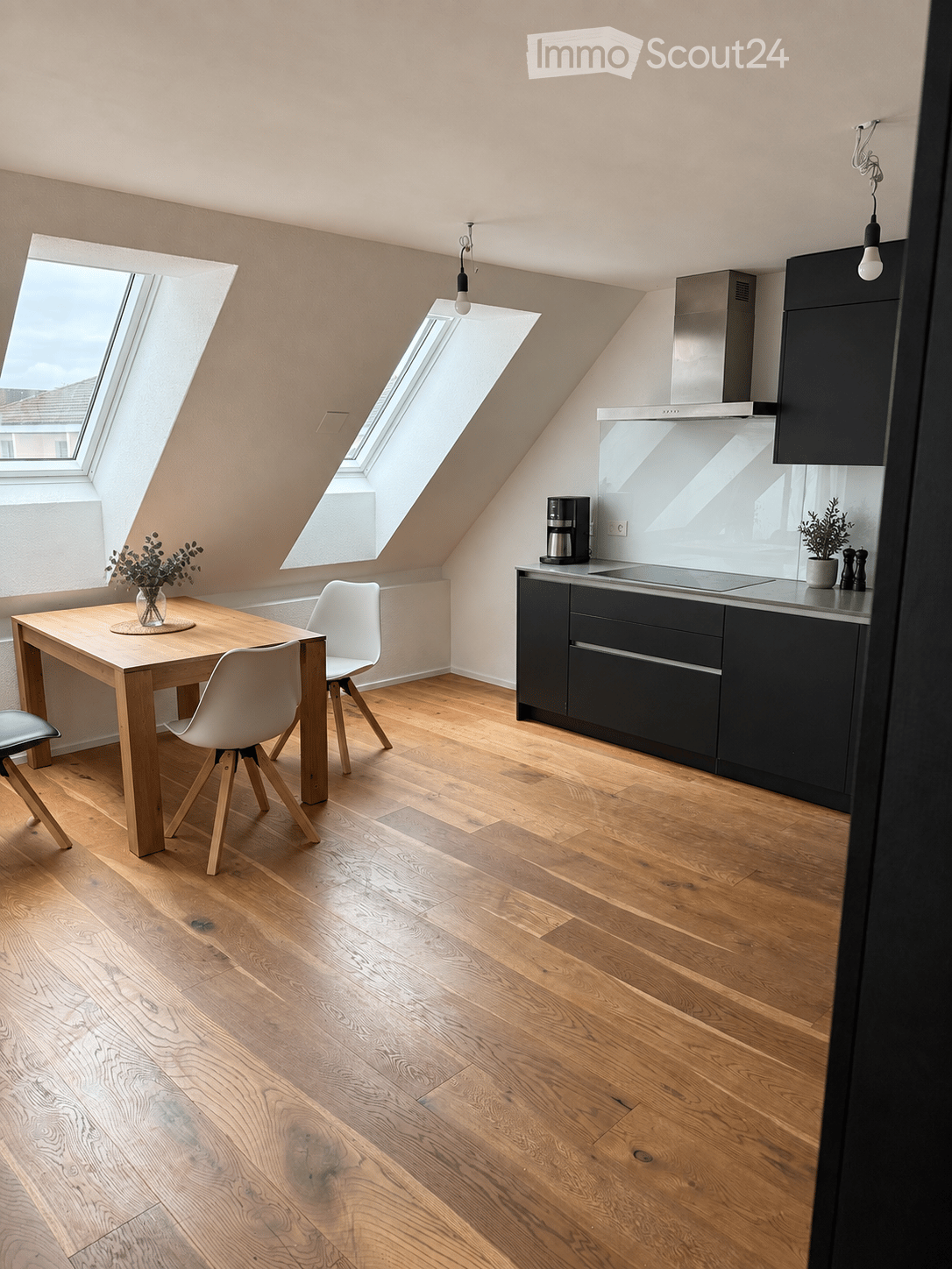 Modern kitchen with wooden flooring, black cabinets, a coffee maker, a small dining table with chairs, and a potted plant on the windowsill.
