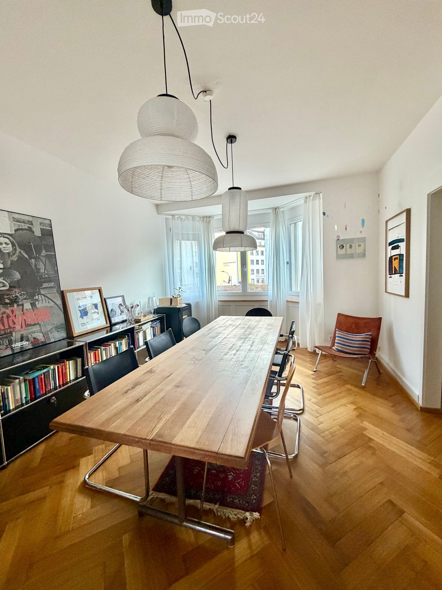 Bright dining area with a long wooden table, multiple chairs, a bookshelf, and modern pendant lights.