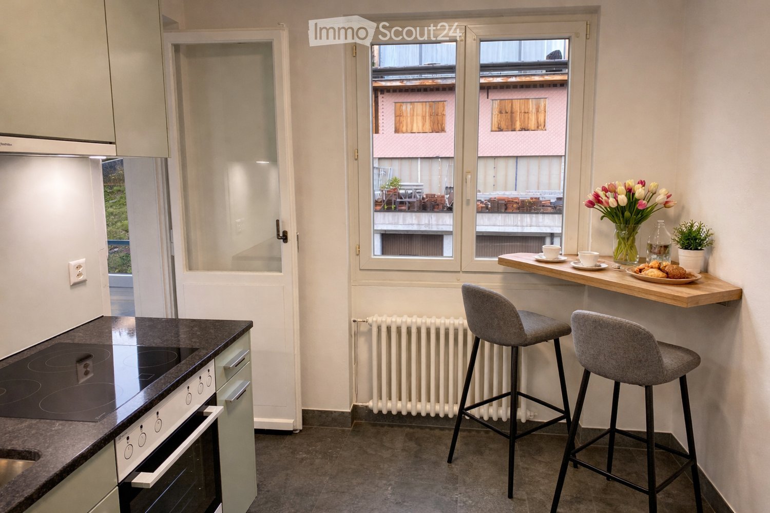 Modern kitchen with an induction stove, oven, bar counter, and two bar stools. A vase with tulips, a cup, a bottle, and a plate with pastries are on the counter. A potted plant is also visible.