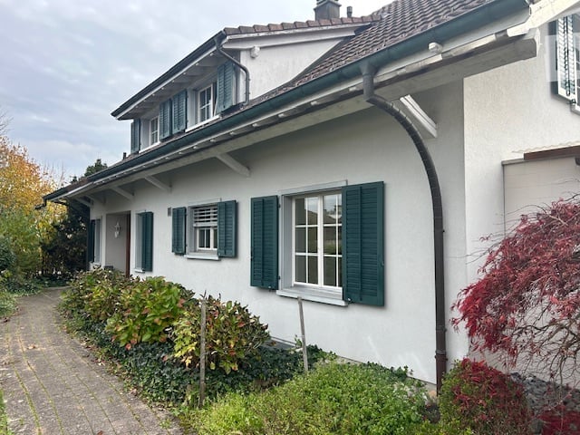 Two-story house with white exterior, green shutters, and multiple windows. The house has a sloped roof and is surrounded by a well-maintained garden with shrubs and trees.
