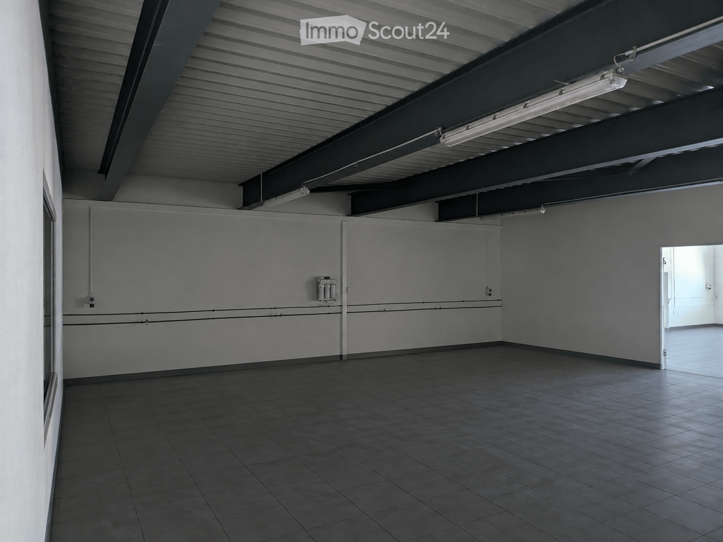 Empty room with tiled floor, white walls, and industrial-style ceiling with exposed beams and fluorescent lighting. No furniture or equipment present.