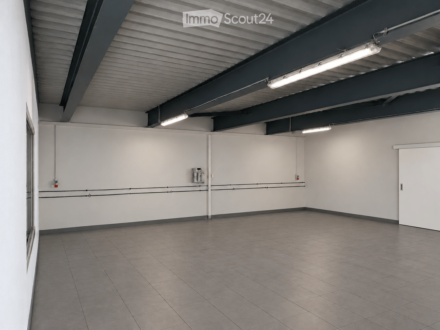 Empty room with tiled floor, white walls, and exposed ceiling beams with fluorescent lighting. No furniture or equipment present.