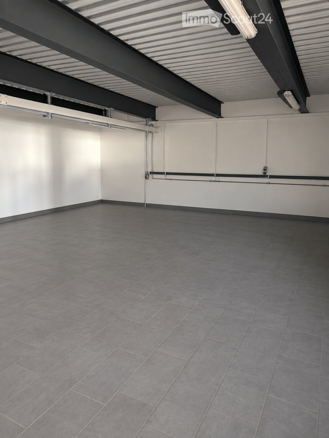 Empty room with grey tiled floor and white walls, featuring exposed metal beams and lighting fixtures on the ceiling.