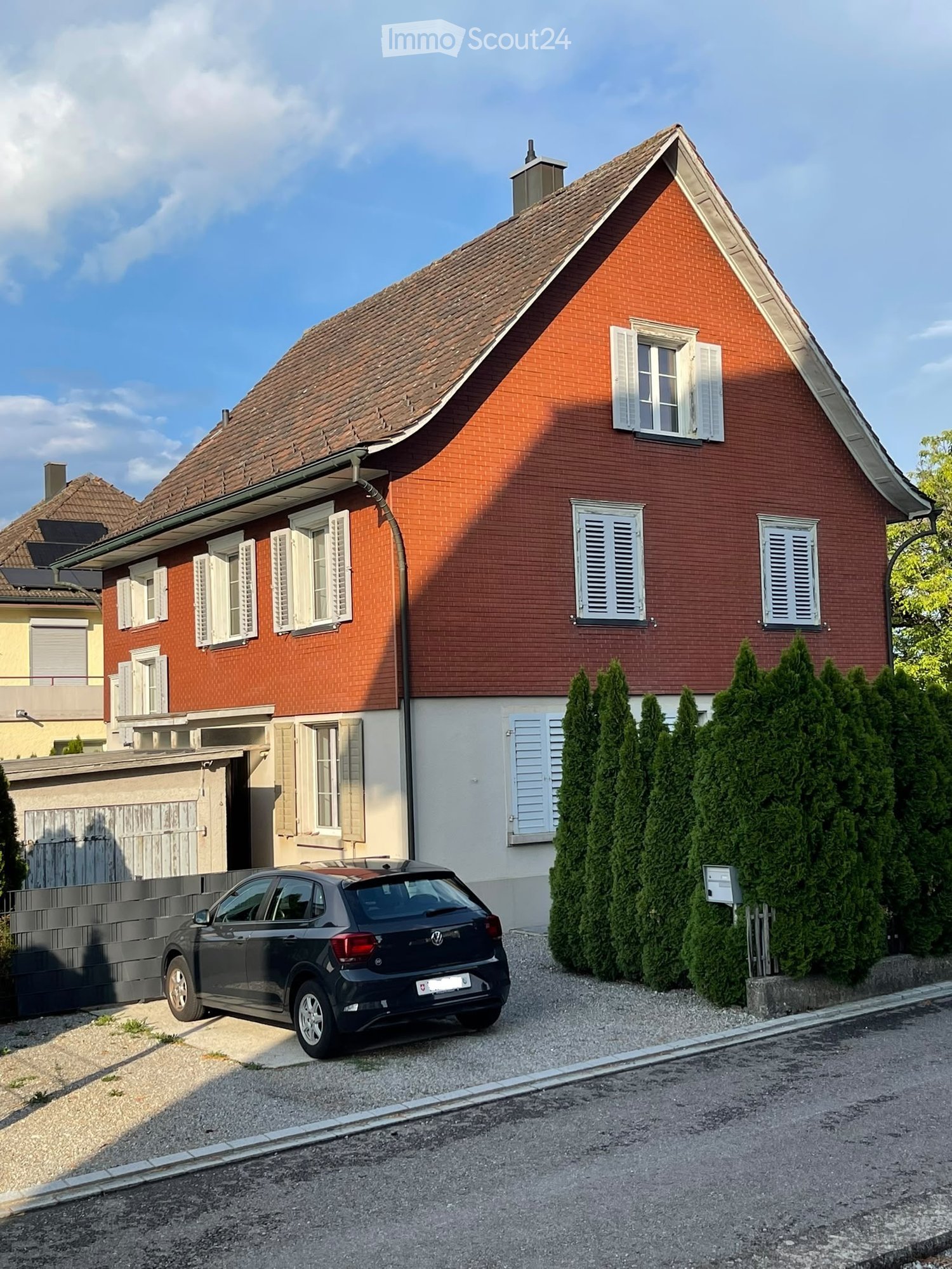 Two-story house with red brick exterior, white shutters, and a tiled roof. A car is parked in the driveway.
