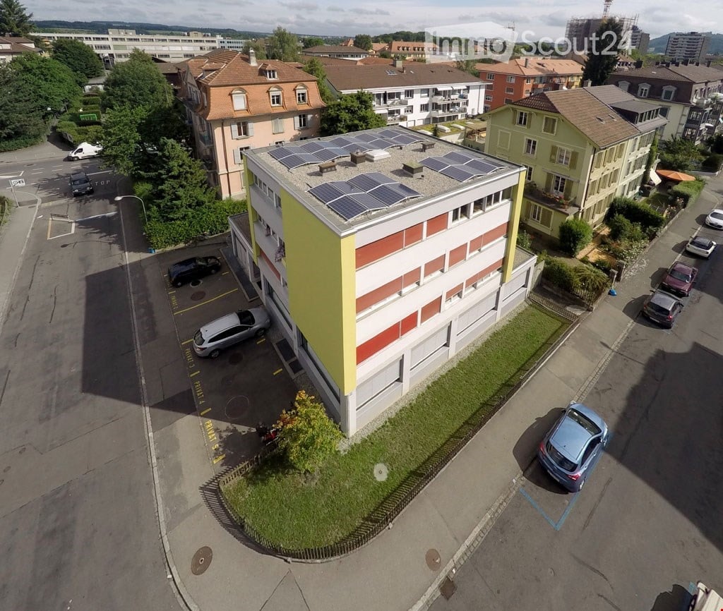 A modern multi-story building with solar panels on the roof, surrounded by parked cars and greenery.