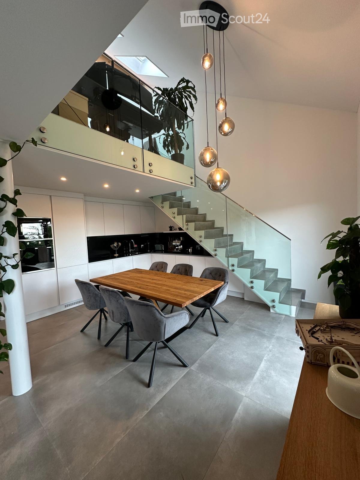 Modern open-plan kitchen and dining area with grey chairs, wooden dining table, white cabinets, and a sleek staircase leading to an upper level.