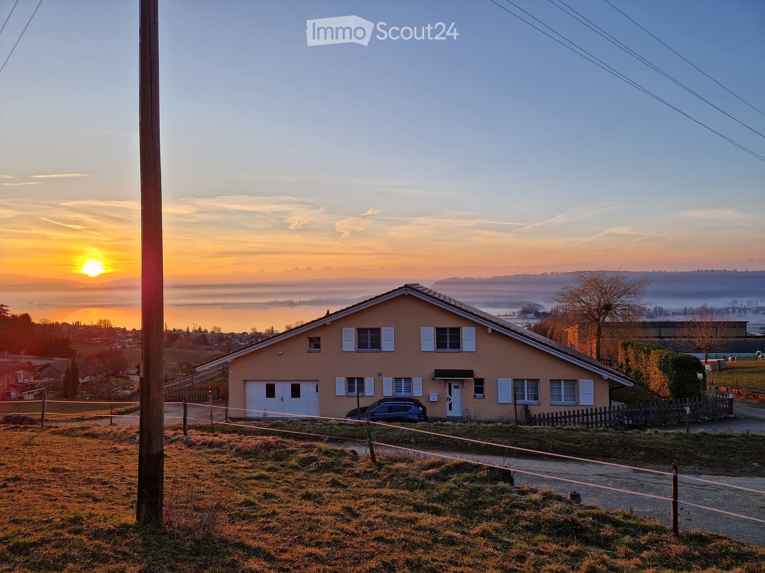 Single-story house with beige walls, white shutters, and a garage. The house is surrounded by a grassy area and a wooden fence, with a scenic view of a lake and distant mountains at sunset.
