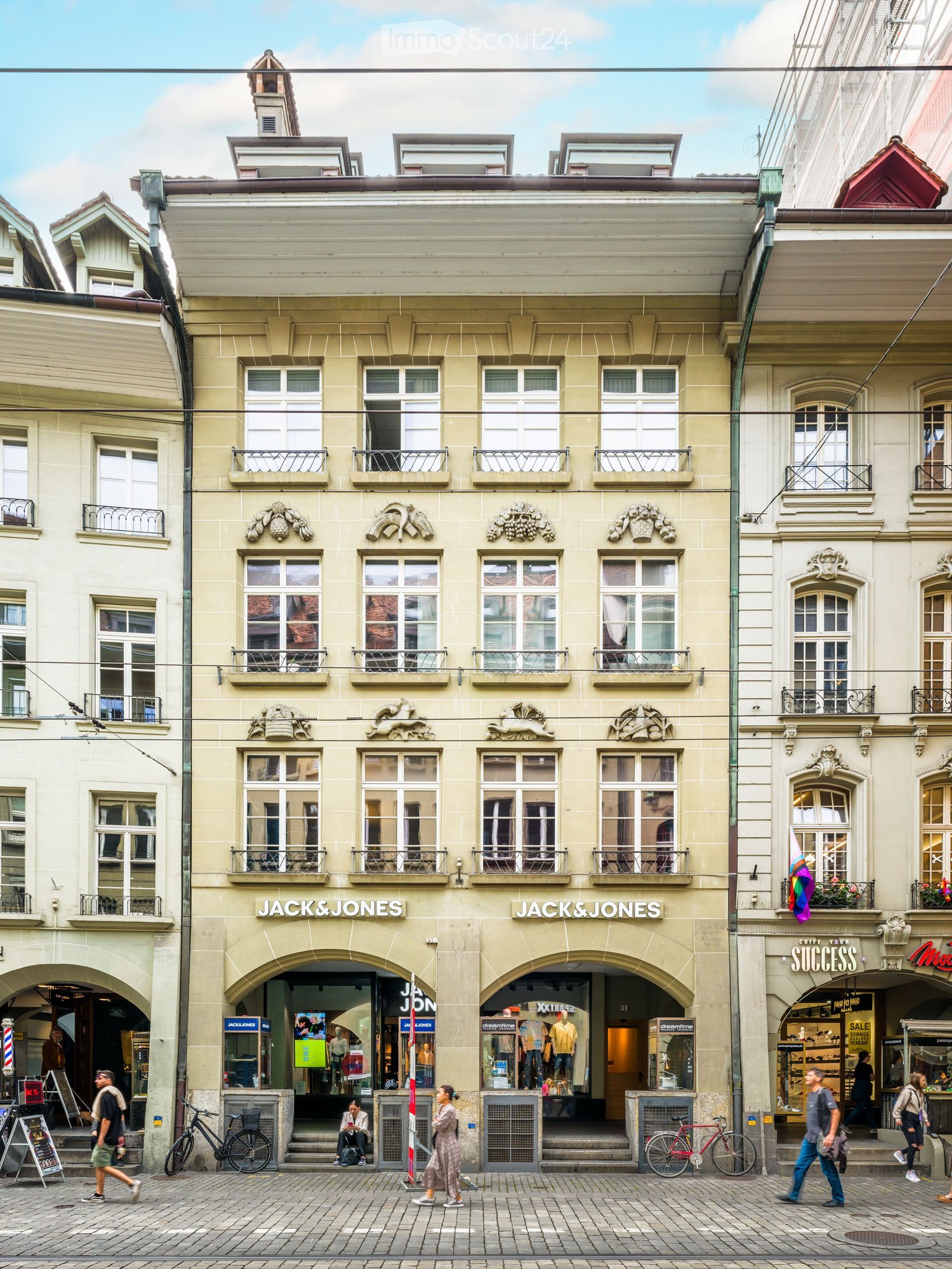 A multi-story building with ornate architectural details, multiple windows, balconies, and storefronts including a Jack & Jones clothing store. The building has a light-colored facade with decorative elements and is situated on a cobblestone street with p
