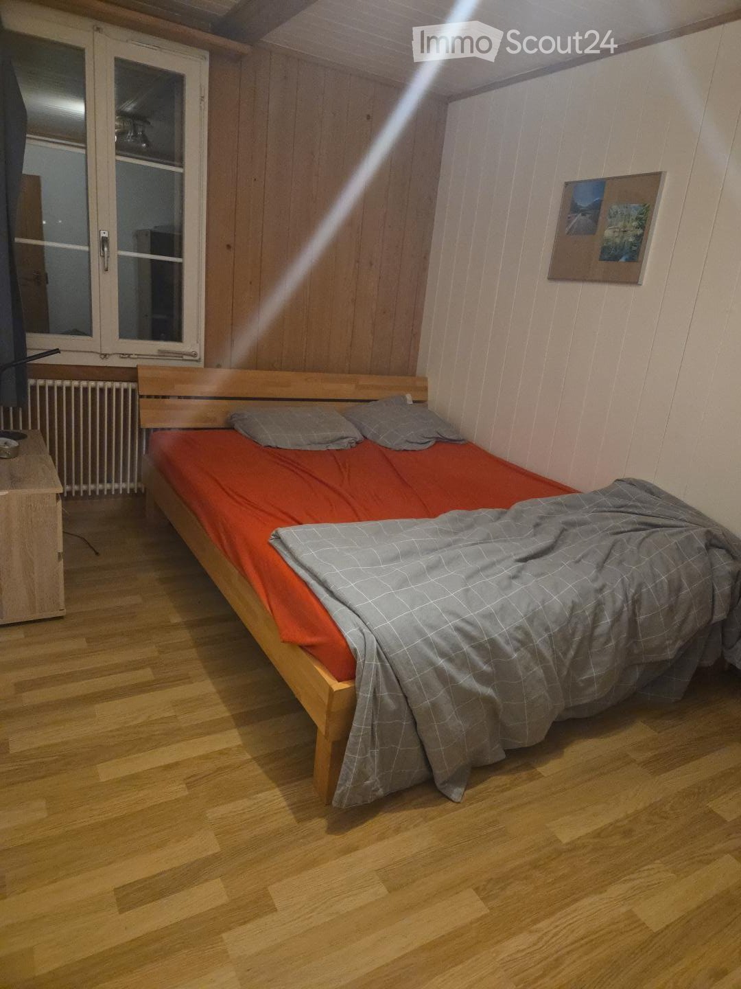 Cozy bedroom with wooden flooring, a single bed with red and grey bedding, a nightstand, radiator, and a picture frame on the wall.