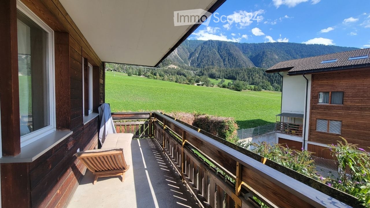 Balcony with wooden railing, overlooking a green field and mountains, with a wooden chair and small table.