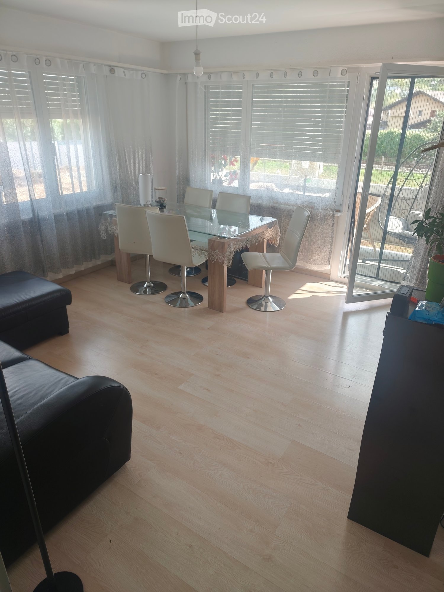 Bright living and dining area with light wooden flooring, large windows with blinds, a black leather sofa, a glass dining table with four white chairs, and a small decorative table runner.