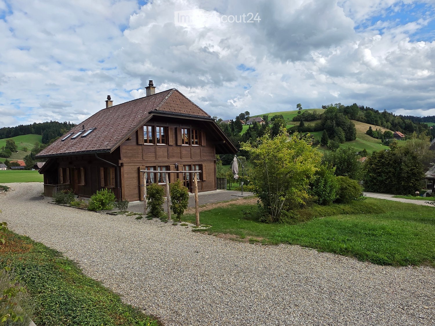 A two-story wooden house with a brown tiled roof, multiple windows, and a chimney, situated in a rural landscape with green hills and trees.