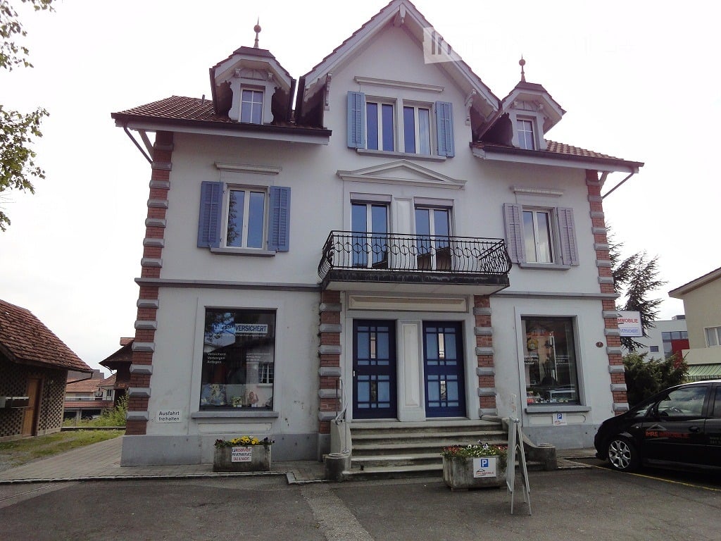 Two-story building with white walls, blue shutters, and a balcony on the second floor. The ground floor has a storefront with glass windows and a door, flanked by flower boxes. The roof features decorative dormers.