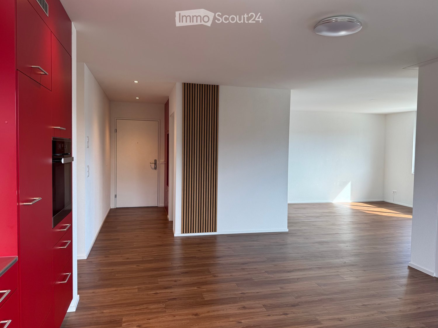 Empty hallway with wooden flooring, white walls, and a red cabinet on the left side. Natural light streams in from the right, illuminating the space.