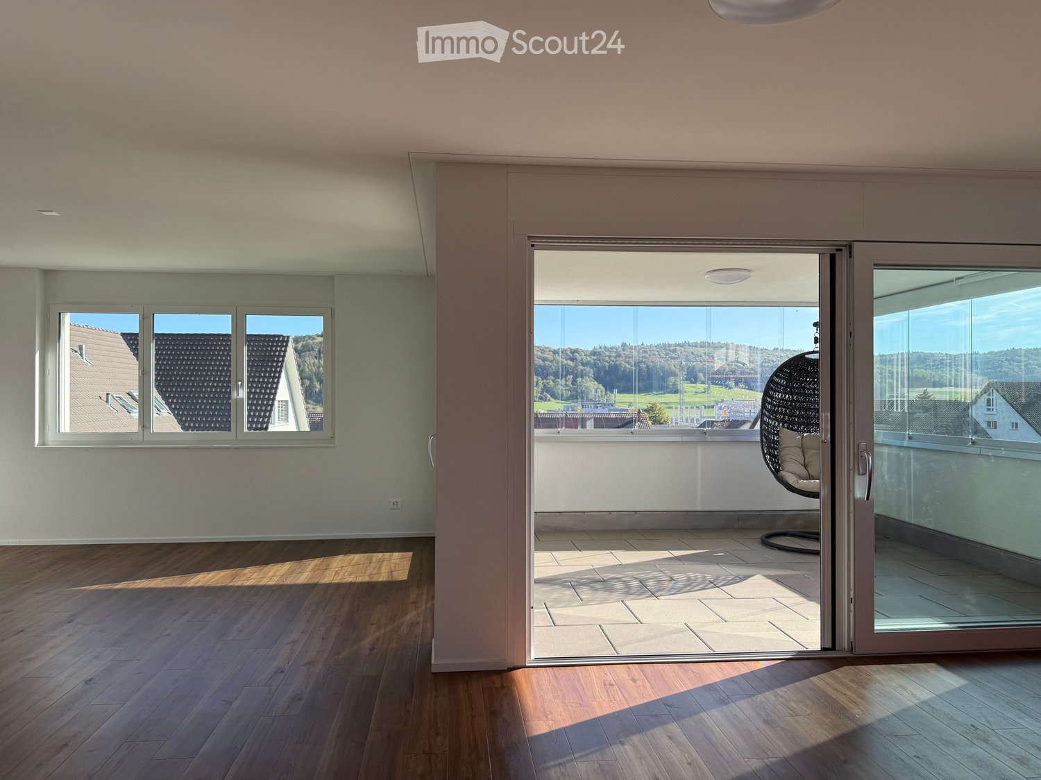 Bright, empty room with wooden flooring, large windows, and a sliding glass door leading to a balcony area with a hanging chair visible outside.