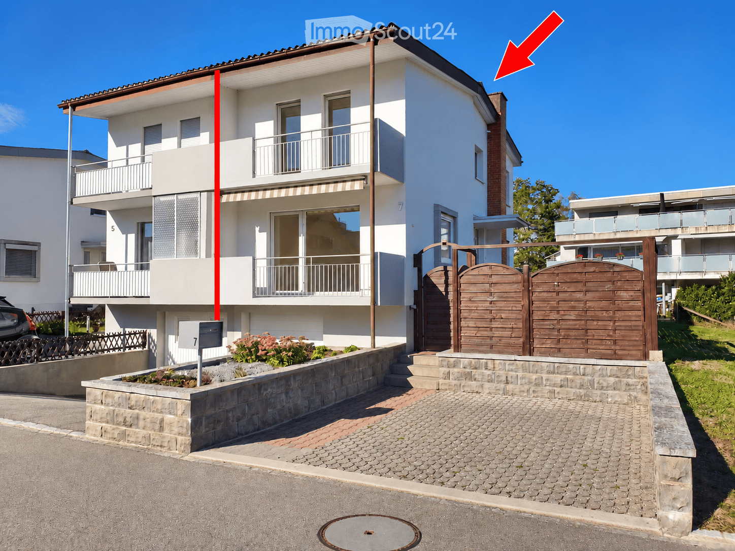 Two-story modern house with white and gray exterior, large windows, balconies, and a wooden gate entrance.