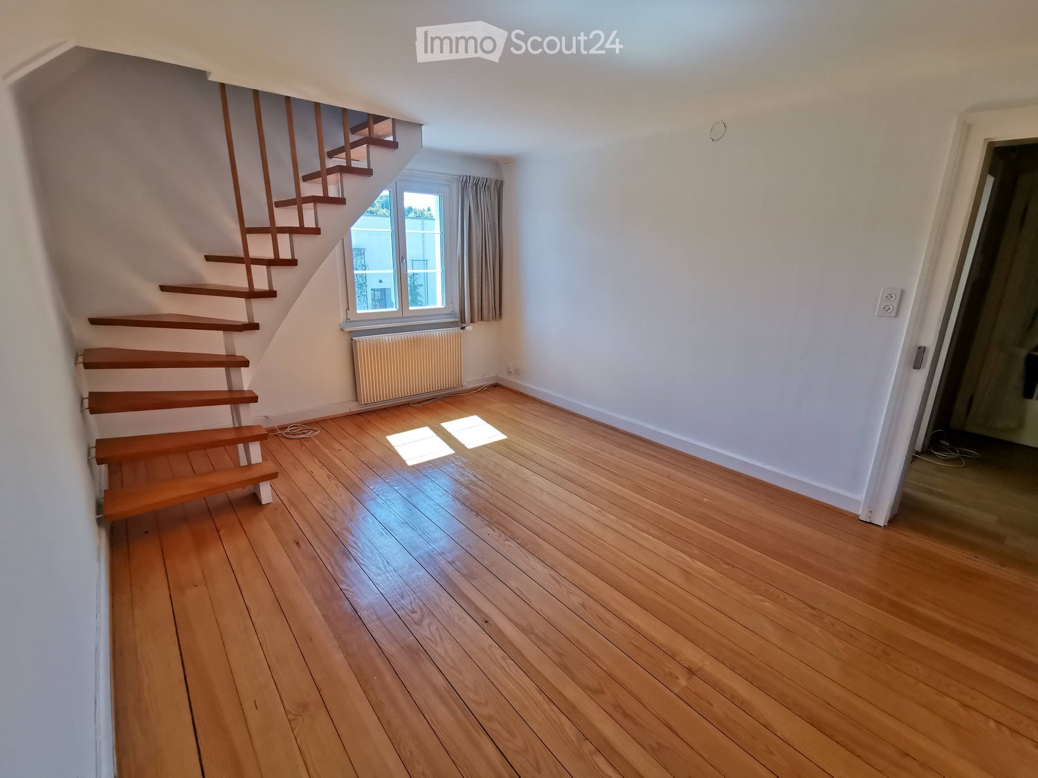 Empty room with wooden flooring, white walls, and a staircase leading upwards. A window with a curtain and a radiator are visible.