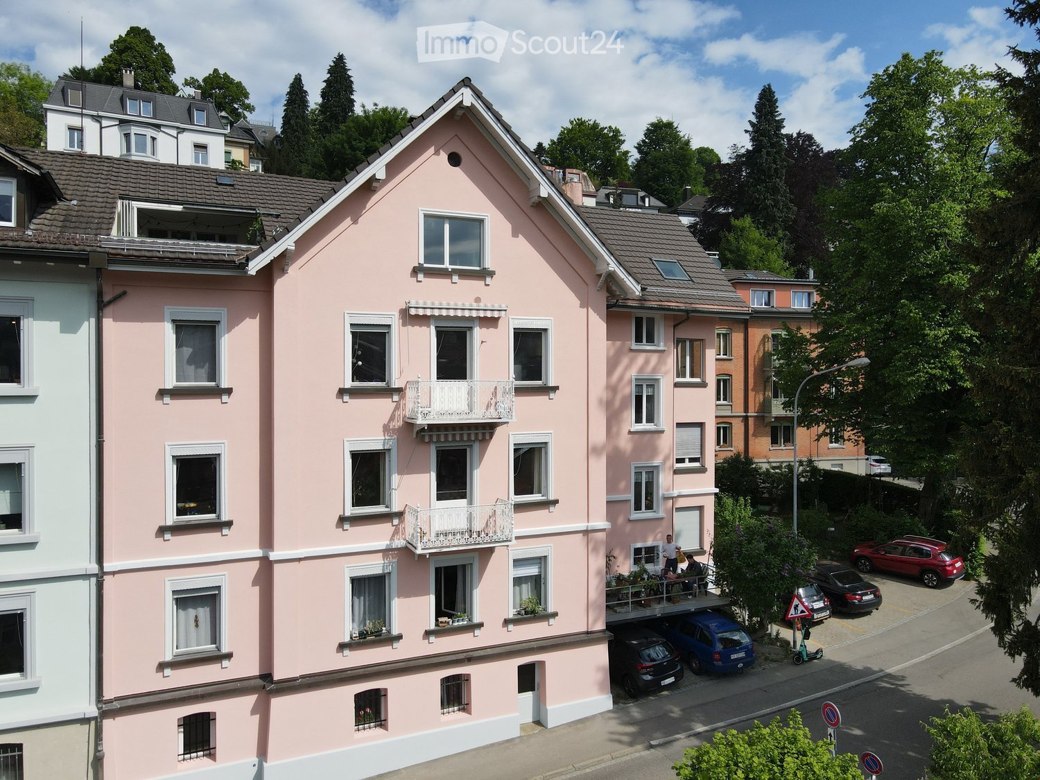 A multi-story pink residential building with white-framed windows, balconies, and a gray tiled roof. The building is surrounded by trees and other houses, with several parked cars visible in the foreground.