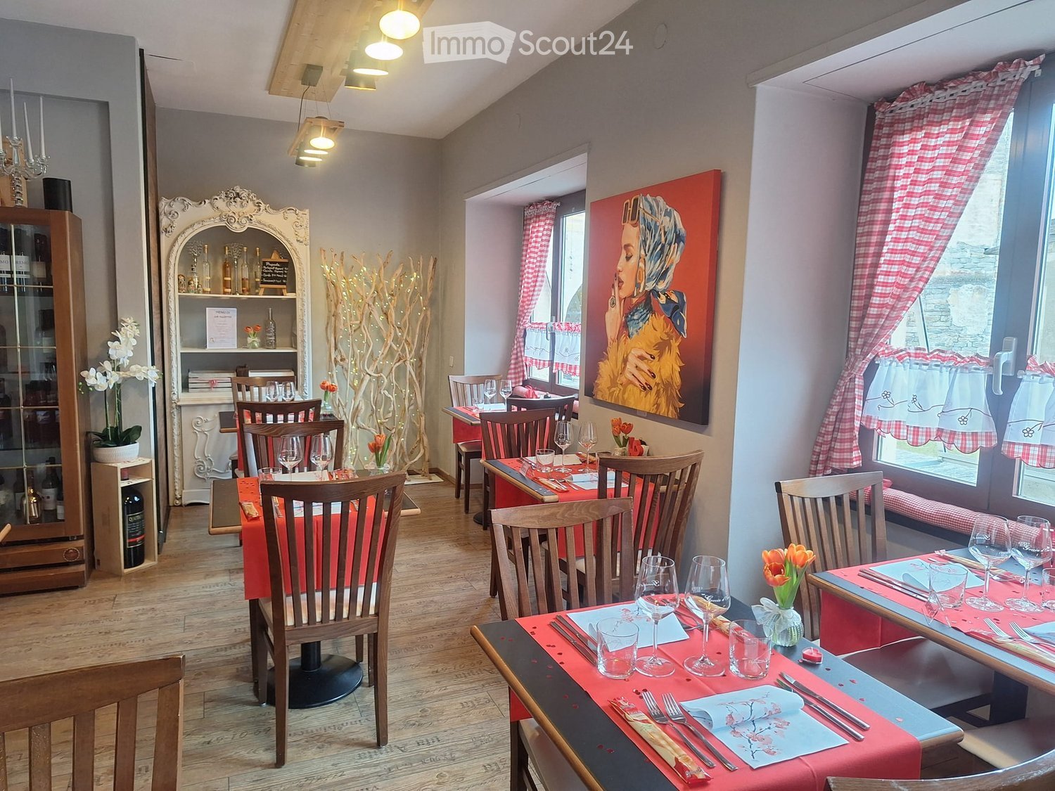 Cozy restaurant interior with wooden tables and chairs, red tablecloths, wine glasses, and decorative elements like a chandelier and artwork.