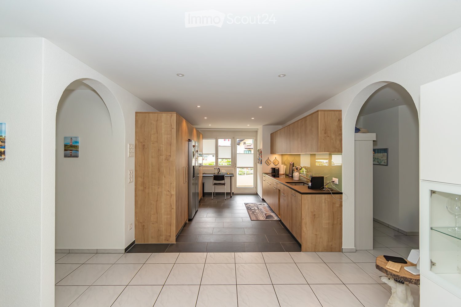 Modern kitchen with light wooden cabinets, stainless steel appliances, and a central island. The space features a tiled floor, recessed lighting, and a small seating area with a table and chairs.