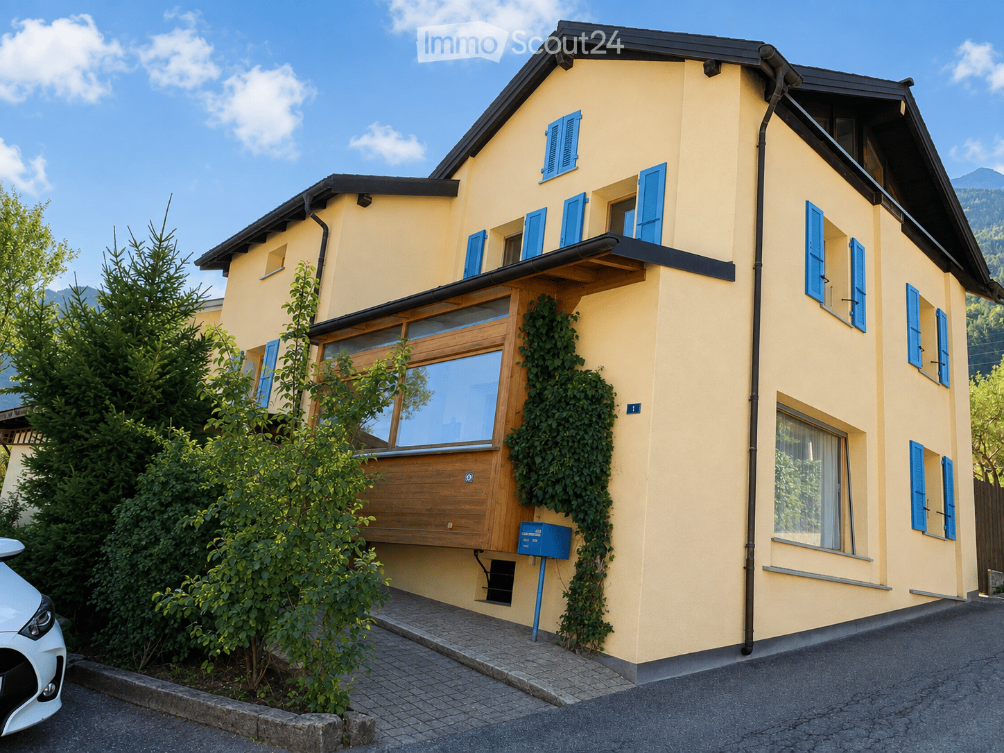 Two-story yellow house with blue shutters, wooden front door, and ivy-covered wall. The house features a balcony and is surrounded by greenery and a parked car.