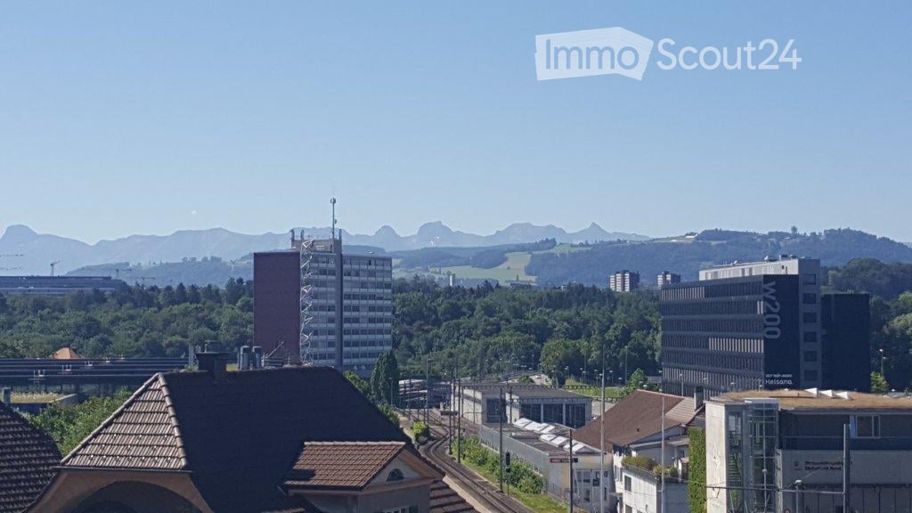 A panoramic view of a cityscape with a mix of modern and traditional buildings, surrounded by green fields and distant mountains under a clear blue sky.