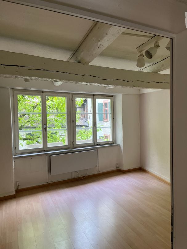 Empty room with wooden flooring, large windows with white frames, radiator, and exposed ceiling beams.