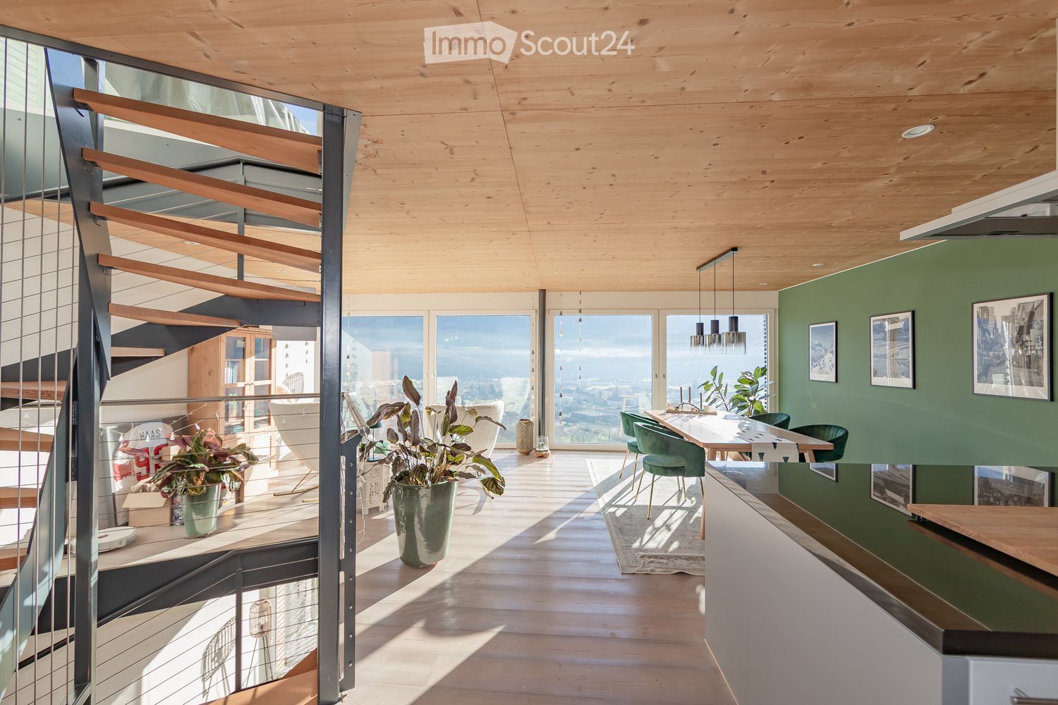 Open-plan living and dining area with wooden floors, large windows offering a city view, a dining table with chairs, potted plants, and a bookshelf.