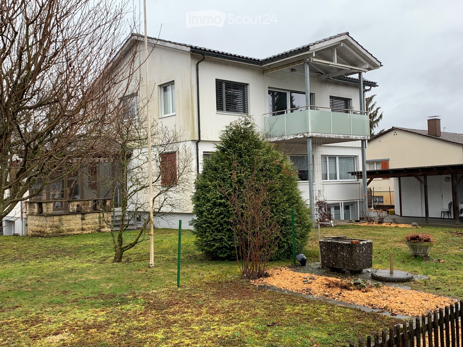 Two-story house with white exterior, multiple windows, raised ground floor, and a balcony on the upper level.