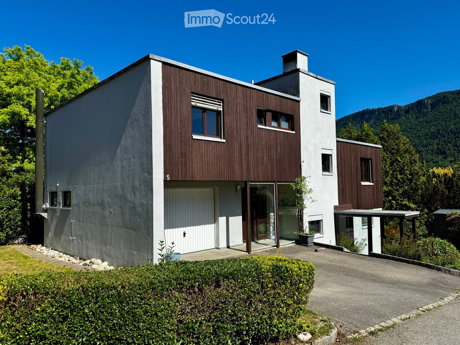 Modern two-story house with white and brown exterior, large windows, and a small garden area.
