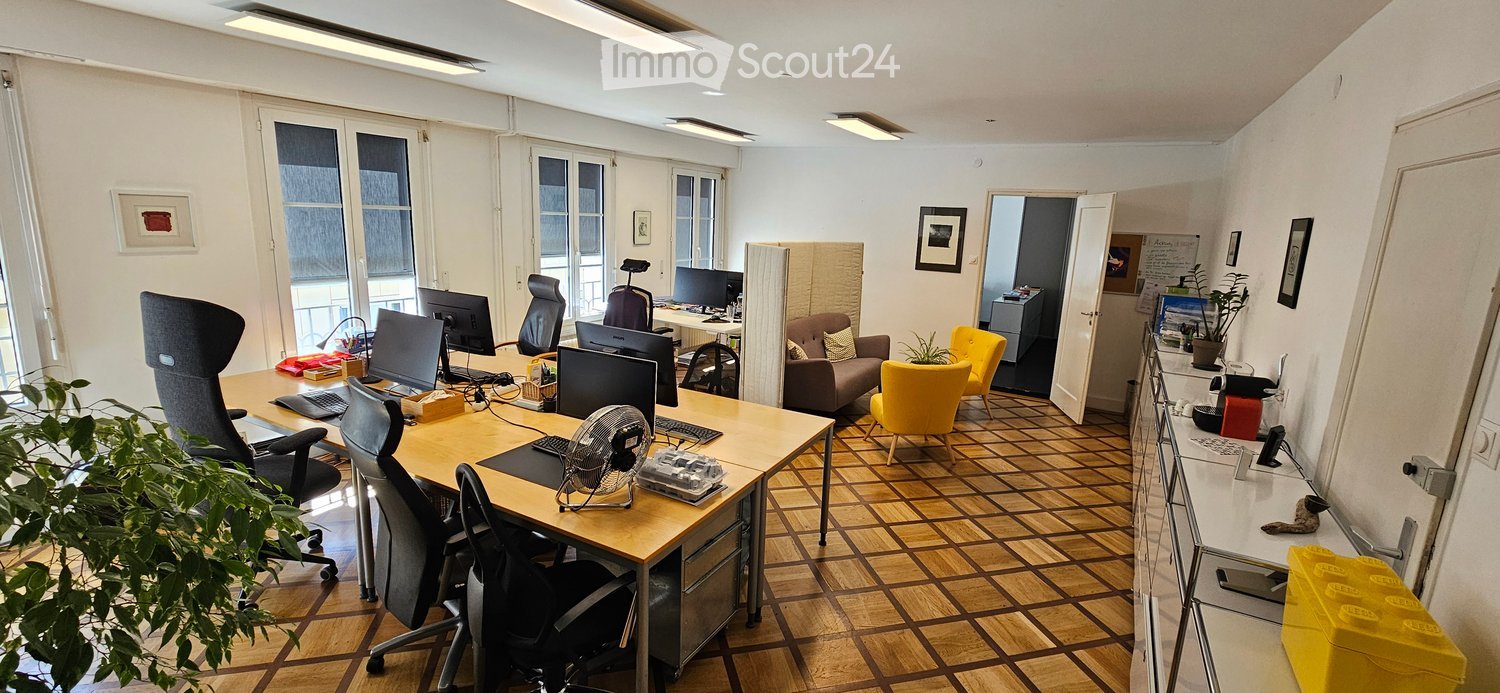 Office space with desks, chairs, computers, a small fan, a sofa, and plants. Large windows provide natural light.