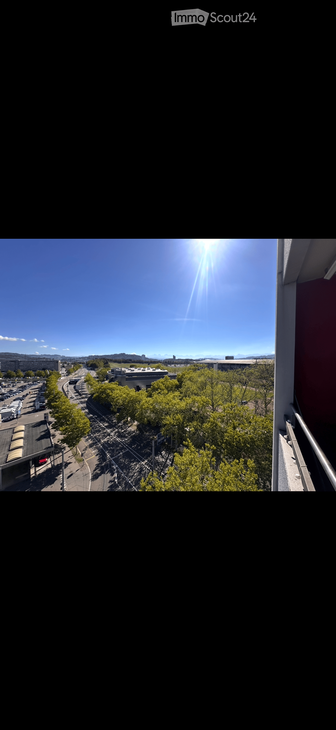 Balcony overlooking a cityscape with trees, parking lot, and distant buildings under a clear blue sky.