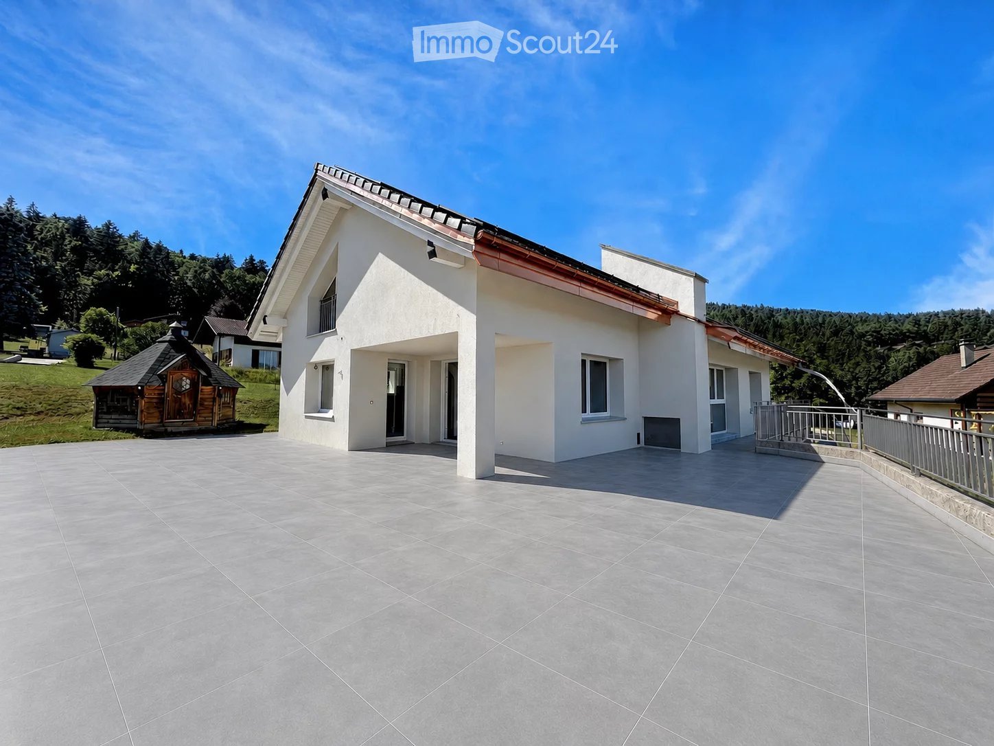A modern two-story house with white walls, large windows, and a tiled patio area. The house features a balcony and is surrounded by greenery and a wooden gazebo.
